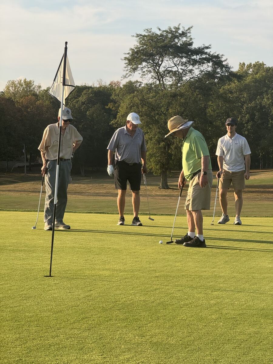 Four golfers on the putting green in the evening light, one lining up a putt beside the flagstick